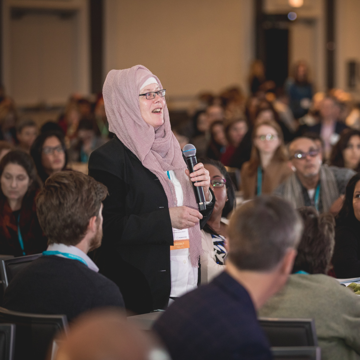 A person with lived experience speaks into a microphone standing among many people sitting at a conference